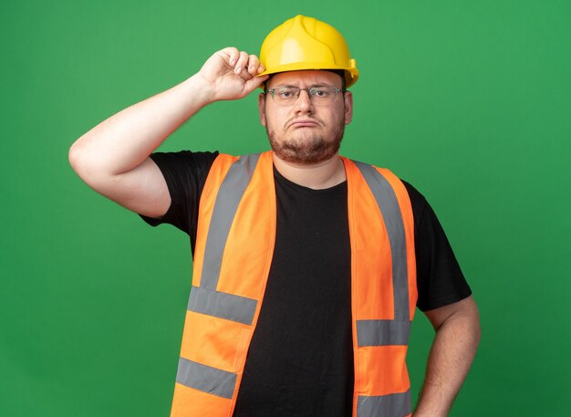 Hombre constructor en chaleco de construcción y casco de seguridad mirando a cámara con rostro serio tocando su casco de pie sobre fondo verde
