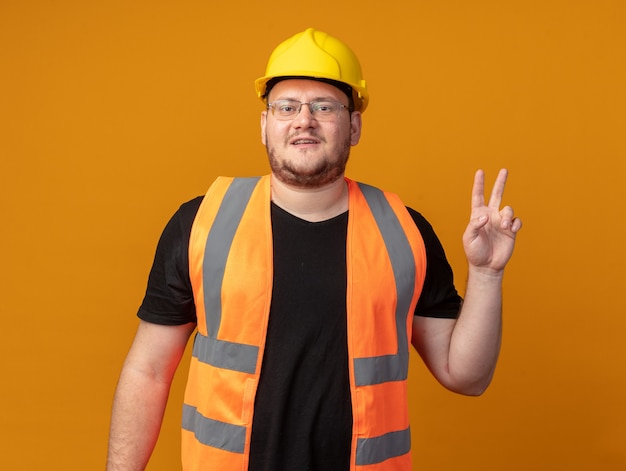 Hombre constructor en chaleco de construcción y casco de seguridad mirando a la cámara sonriendo confiado mostrando v-sign de pie sobre fondo naranja