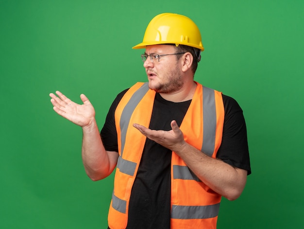 Hombre constructor en chaleco de construcción y casco de seguridad mirando a un lado confundido levantando los brazos con indignación