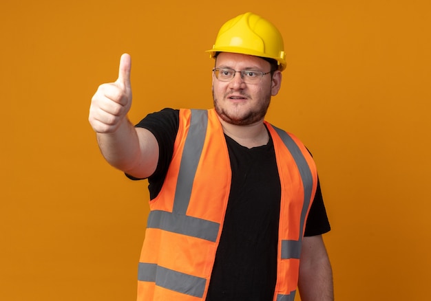 Hombre constructor en chaleco de construcción y casco de seguridad mirando a un lado sonriendo confiado mostrando los pulgares para arriba sobre fondo naranja