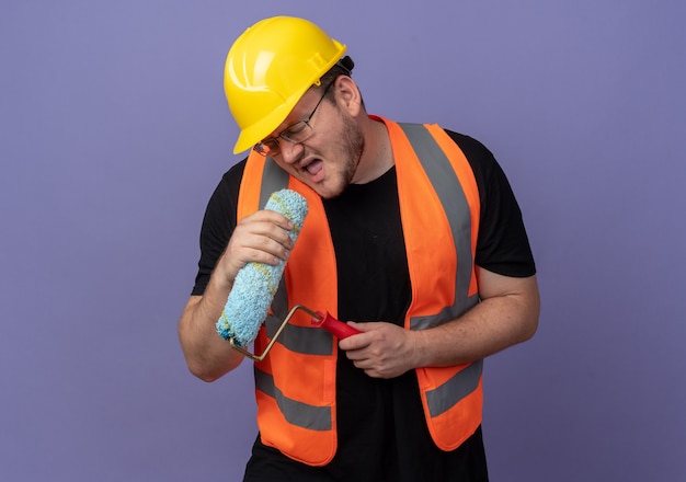 Hombre constructor en chaleco de construcción y casco de seguridad con rodillo de pintura usándolo como micrófono cantando divirtiéndose