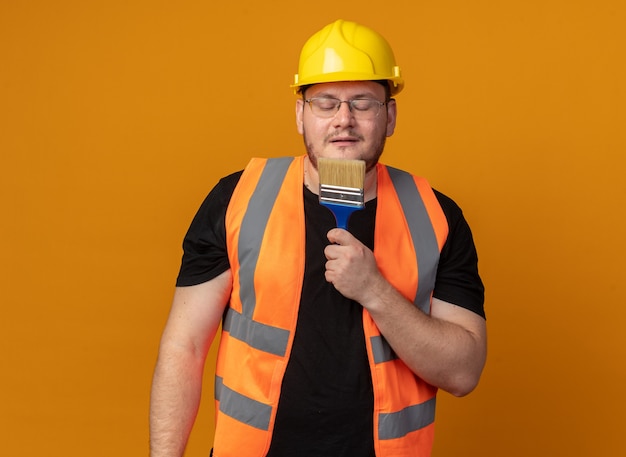 Hombre constructor en chaleco de construcción y casco de seguridad sosteniendo pincel usándolo como un micrófono cantando