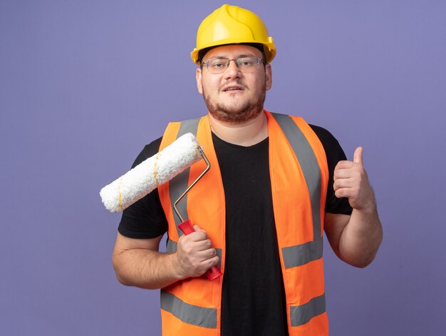 Hombre constructor en chaleco de construcción y casco de seguridad sosteniendo el rodillo de pintura mirando a la cámara sonriendo confiado mostrando los pulgares para arriba sobre fondo azul.