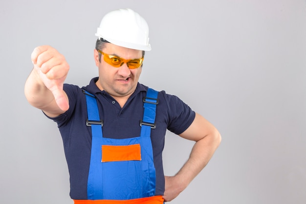 Foto gratuita hombre constructor disgustado de mediana edad vistiendo uniforme de construcción y casco de seguridad mostrando los pulgares hacia abajo sobre la pared blanca aislada