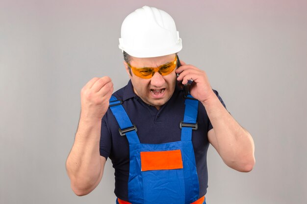 Hombre constructor enojado de mediana edad vistiendo uniforme de construcción y casco de seguridad hablando por teléfono móvil gritando con ira loco y gritando con la mano levantada sobre la pared blanca aislada