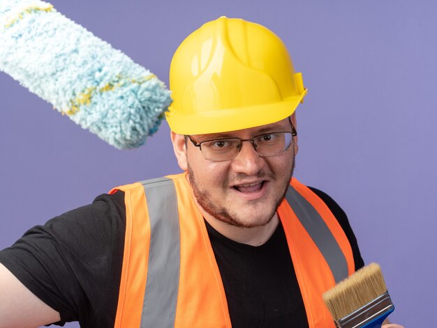 Hombre constructor feliz en chaleco de construcción y casco de seguridad con rodillo de pintura y pincel