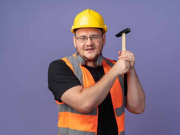 Hombre constructor feliz en chaleco de construcción y casco de seguridad sosteniendo un martillo mirando a la cámara sonriendo alegremente de pie sobre azul