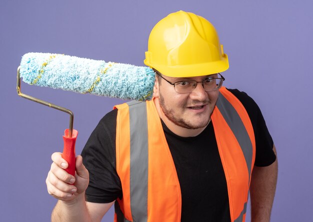 Hombre constructor feliz en chaleco de construcción y casco de seguridad sosteniendo el rodillo de pintura mirando a la cámara sonriendo alegremente de pie sobre azul