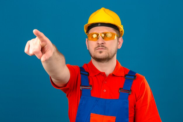 Hombre constructor con gafas de uniforme de construcción y casco de seguridad apuntando con el dedo a algo delante de él con cara seria de pie sobre la pared azul aislada