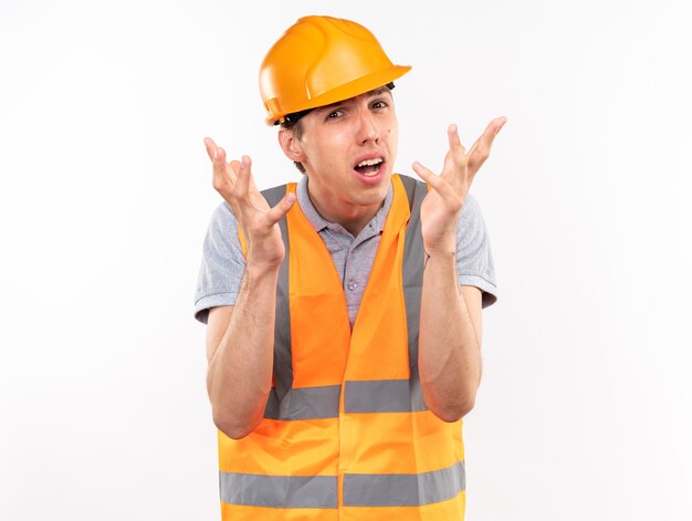Hombre constructor joven confundido en uniforme extendiendo las manos aisladas en la pared blanca