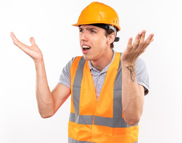 Hombre constructor joven confundido en uniforme extendiendo las manos aisladas en la pared blanca