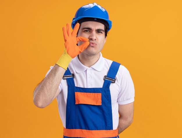 Hombre constructor joven enojado en uniforme de construcción y casco de seguridad con guantes de goma haciendo gesto de silencio como cerrar la boca con una cremallera de pie sobre una pared naranja