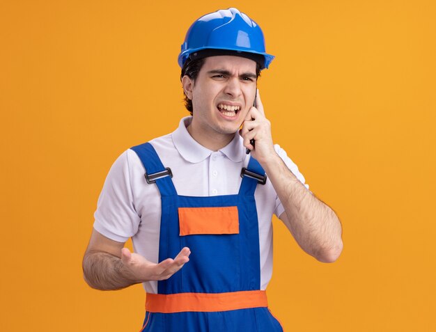 Hombre constructor joven enojado en uniforme de construcción y casco de seguridad hablando por teléfono móvil de pie sobre la pared naranja