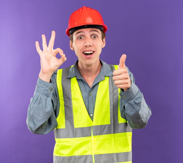 Hombre constructor joven sorprendido en uniforme mostrando gesto bien con el pulgar hacia arriba