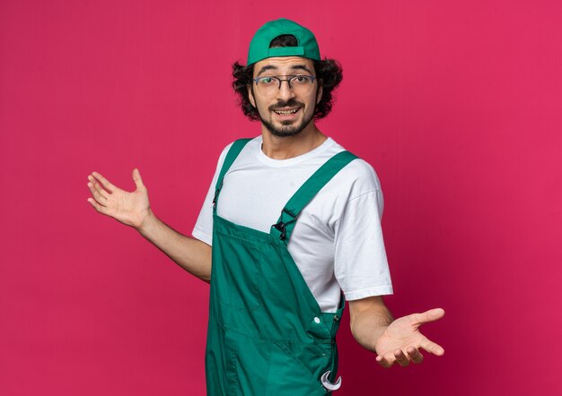 Hombre constructor joven sorprendido vestido con uniforme con tapa extendiendo las manos