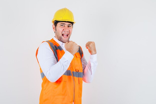 Hombre constructor mostrando gesto ganador en camisa, uniforme y mirando feliz. vista frontal.