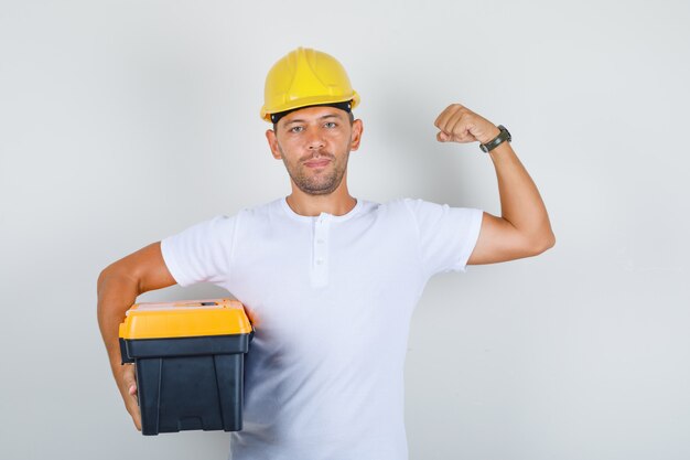 Hombre constructor sosteniendo caja de herramientas y mostrando músculo en camiseta, casco y mirando confiado, vista frontal.