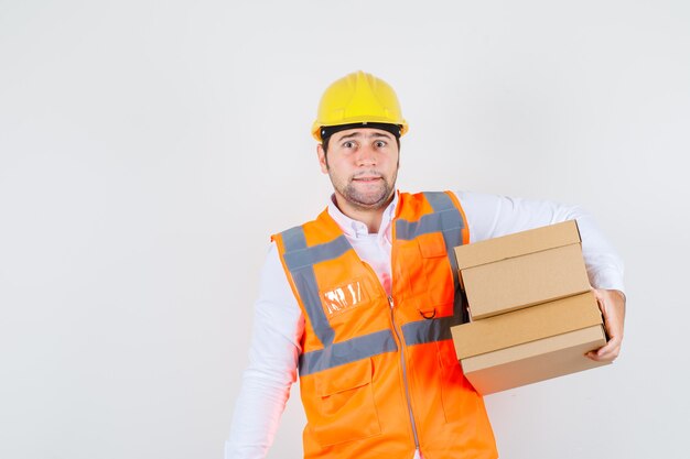 Hombre constructor sosteniendo cajas de cartón en camisa, uniforme y mirando preocupado, vista frontal.