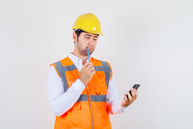 Hombre constructor sosteniendo un lápiz mientras mira el teléfono inteligente en camisa, uniforme y mirando pensativo, vista frontal.