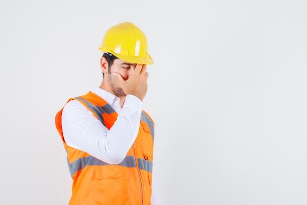 Hombre constructor sosteniendo la mano en la cara en camisa, uniforme y mirando exhausto. vista frontal.