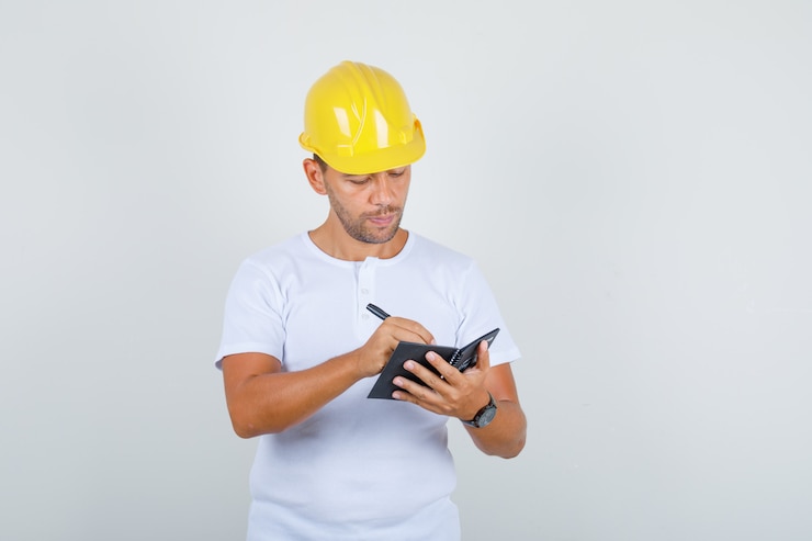 Foto gratuito hombre constructor tomando notas en el mini portátil en camiseta blanca, casco y mirando ocupado, vista frontal