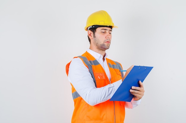 Hombre constructor tomando notas en el portapapeles en camisa, uniforme y mirando ocupado, vista frontal.