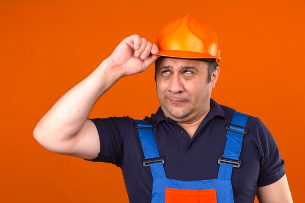Hombre constructor con uniforme de construcción y casco de seguridad con cara de tristeza y decepción al tocar su casco sobre una pared naranja aislada
