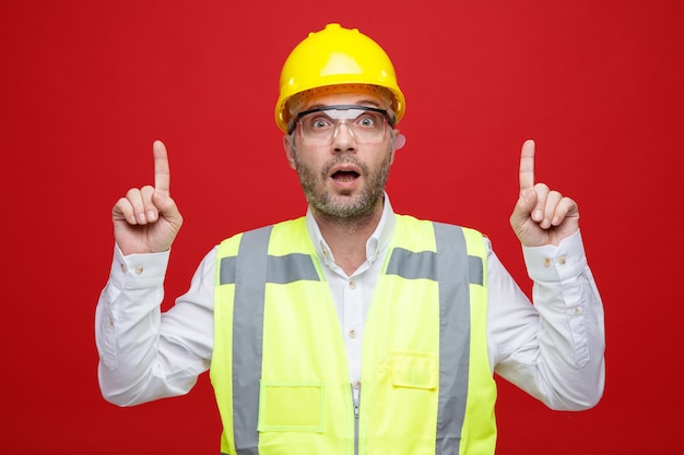 Foto gratuita hombre constructor en uniforme de construcción y casco de seguridad con gafas de seguridad mirando a la cámara apuntando con los dedos índices de pie sobre fondo rosa