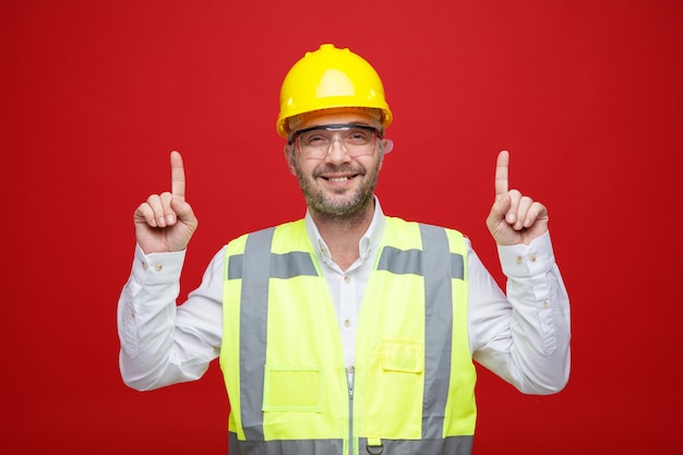 Hombre constructor en uniforme de construcción y casco de seguridad con gafas de seguridad mirando a la cámara feliz y alegre señalando con los dedos índices de pie sobre fondo rosa