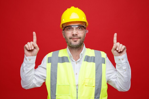 Hombre constructor en uniforme de construcción y casco de seguridad con gafas de seguridad mirando a la cámara feliz y positivo señalando con los dedos índices de pie sobre fondo rosa