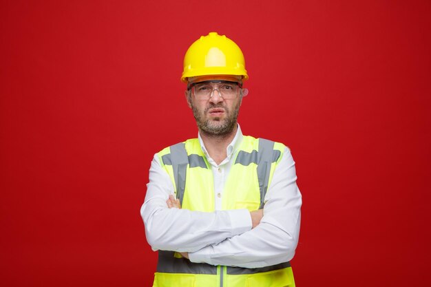 Hombre constructor en uniforme de construcción y casco de seguridad con gafas de seguridad mirando a la cámara insatisfecho de pie sobre fondo rosa