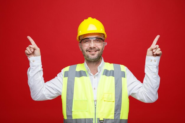 Hombre constructor en uniforme de construcción y casco de seguridad con gafas de seguridad mirando a la cámara sonriendo feliz y positivo señalando con los dedos índices de pie sobre fondo rosa