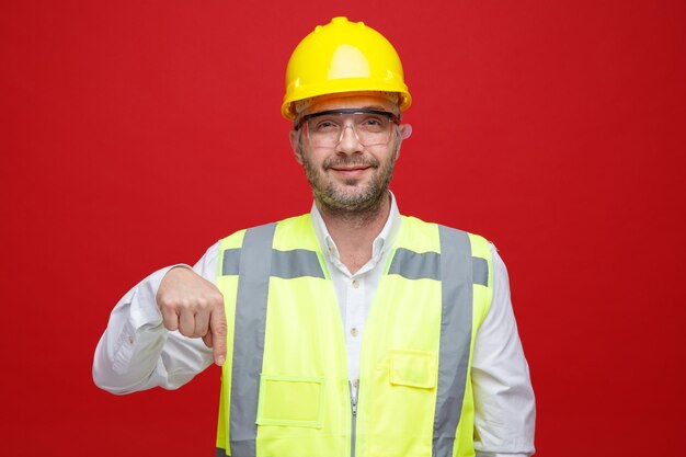 Hombre constructor en uniforme de construcción y casco de seguridad con gafas de seguridad mirando a la cámara sonriendo señalando con el dedo índice hacia abajo de pie sobre fondo rosa