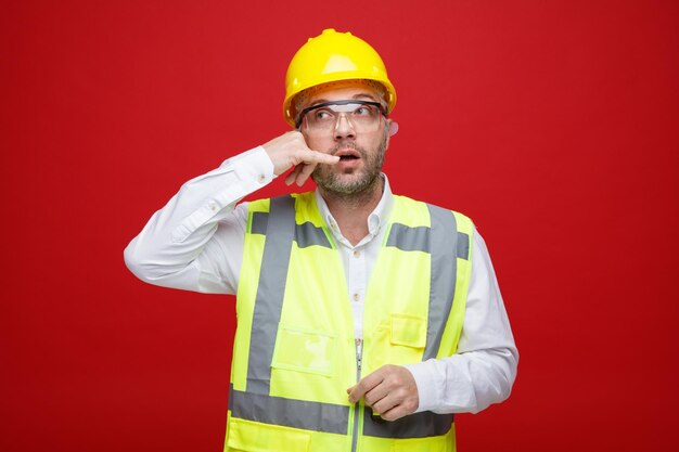 Hombre constructor en uniforme de construcción y casco de seguridad con gafas de seguridad mirando a un lado haciendo un gesto de llamarme de pie sobre fondo rojo.