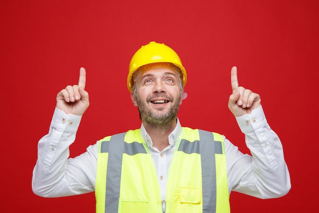 Hombre constructor en uniforme de construcción y casco de seguridad mirando hacia arriba sonriendo alegremente señalando con los dedos índices de pie sobre fondo rojo.