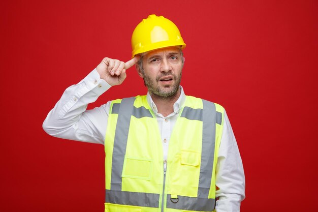 Hombre constructor en uniforme de construcción y casco de seguridad mirando a la cámara desconcertado rascándose la cabeza de pie sobre fondo rojo.