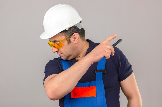 Hombre constructor con uniforme de construcción y casco de seguridad mirando a otro lado y apuntando a algo sobre la pared blanca aislada