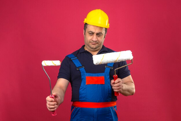 Hombre constructor con uniforme de construcción y casco de seguridad de pie con rodillos de pintura mirándolos sintiéndose escéptico dudoso sobre pared rosa aislado