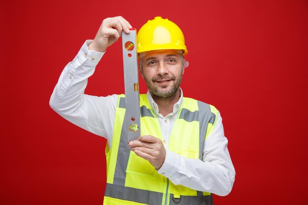 Hombre constructor en uniforme de construcción y casco de seguridad con regla de nivel mirando a la cámara con una sonrisa en la cara feliz de pie sobre fondo rojo.