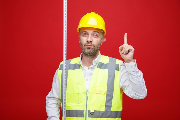 Hombre constructor en uniforme de construcción y casco de seguridad sosteniendo cinta métrica mirando a la cámara con cara seria apuntando con el dedo índice hacia arriba de pie sobre fondo rojo.