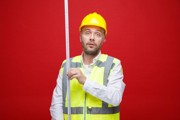 Hombre constructor en uniforme de construcción y casco de seguridad sosteniendo cinta métrica mirando a la cámara con expresión triste en la cara de pie sobre fondo rojo.