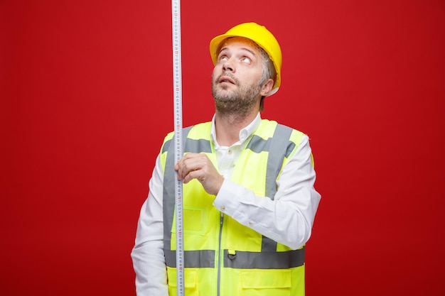 Hombre constructor en uniforme de construcción y casco de seguridad sosteniendo cinta métrica mirando desconcertado de pie sobre fondo rojo.