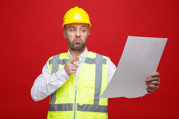 Hombre constructor en uniforme de construcción y casco de seguridad sosteniendo un plan mirándolo confundido de pie sobre fondo rojo.