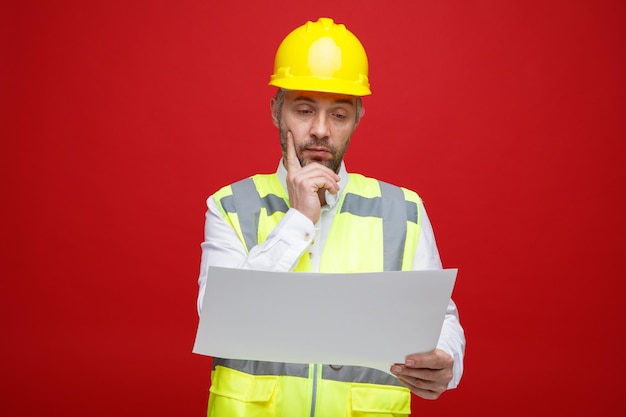 Hombre constructor en uniforme de construcción y casco de seguridad sosteniendo un plan mirándolo con interés pensando de pie sobre fondo rojo.