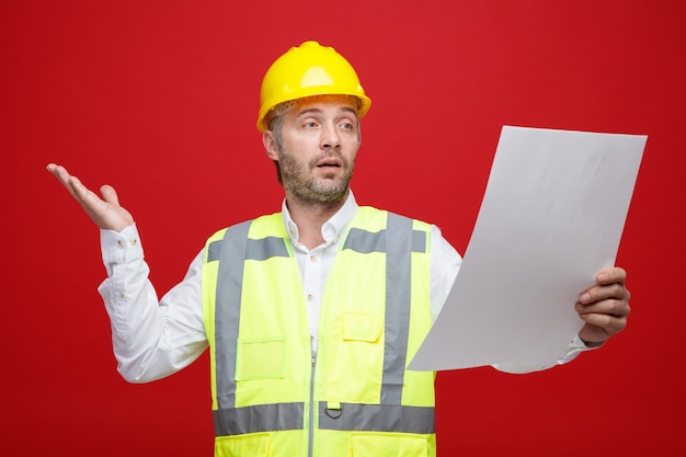 Hombre constructor en uniforme de construcción y casco de seguridad sosteniendo un plan que parece confundido levantando el brazo sin respuesta de pie sobre fondo rojo