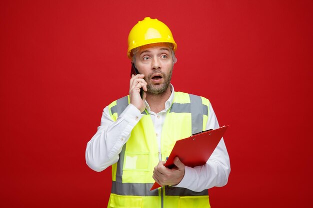 Hombre constructor en uniforme de construcción y casco de seguridad sosteniendo portapapeles hablando por teléfono móvil mirando confundido de pie sobre fondo rojo.