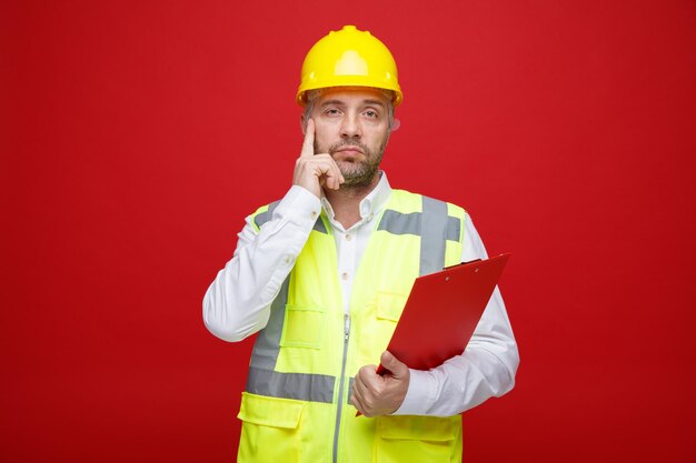 Hombre constructor en uniforme de construcción y casco de seguridad sosteniendo portapapeles mirando a la cámara con expresión pensativa de pie sobre fondo rojo.