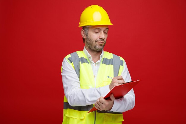 Hombre constructor en uniforme de construcción y casco de seguridad sosteniendo portapapeles mirando confiado haciendo notas de pie sobre fondo rojo.