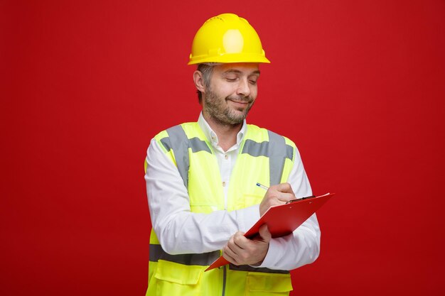 Hombre constructor en uniforme de construcción y casco de seguridad sosteniendo portapapeles mirando confiado haciendo notas de pie sobre fondo rojo.