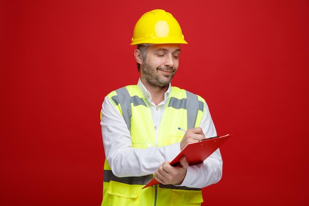 Hombre constructor en uniforme de construcción y casco de seguridad sosteniendo portapapeles mirando confiado haciendo notas de pie sobre fondo rojo.